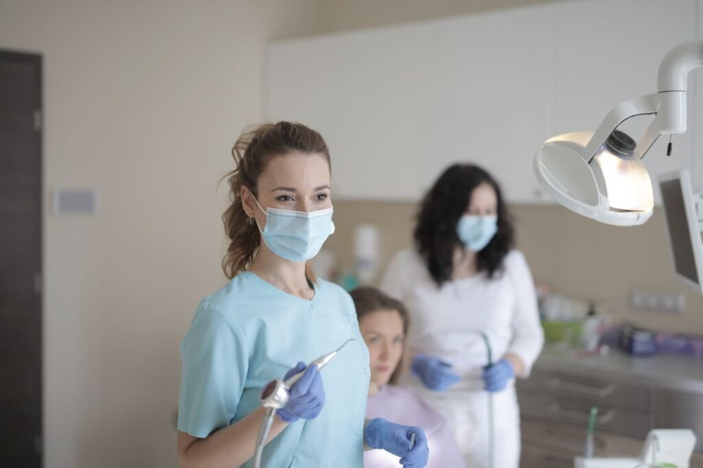 Young female dentist in medical mask and latex gloves using medical instrument while working with colleague during treatment of patient in modern dental room